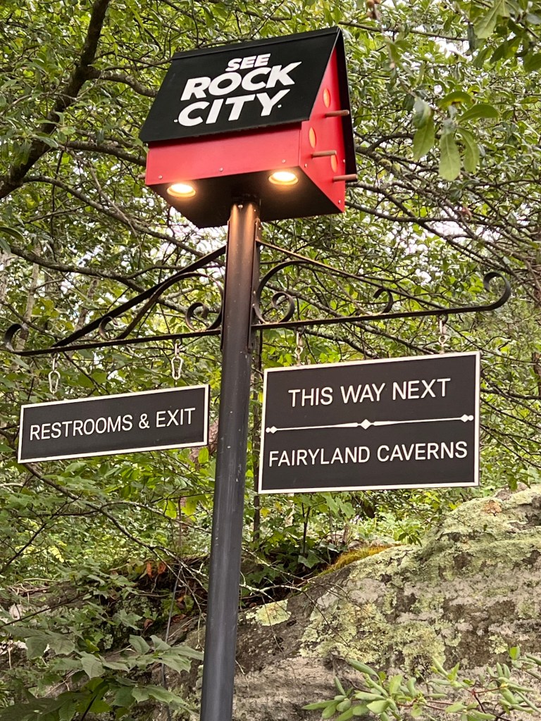 Sign for Rock City with directions to restrooms and Fairyland Caverns, surrounded by greenery.
